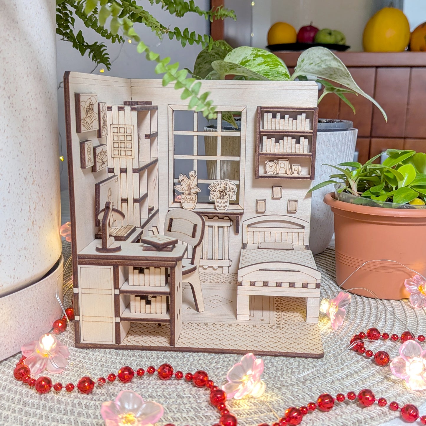 Miniature wooden laser cut model of a room with furniture and decor, surrounded by plants and decorative items.