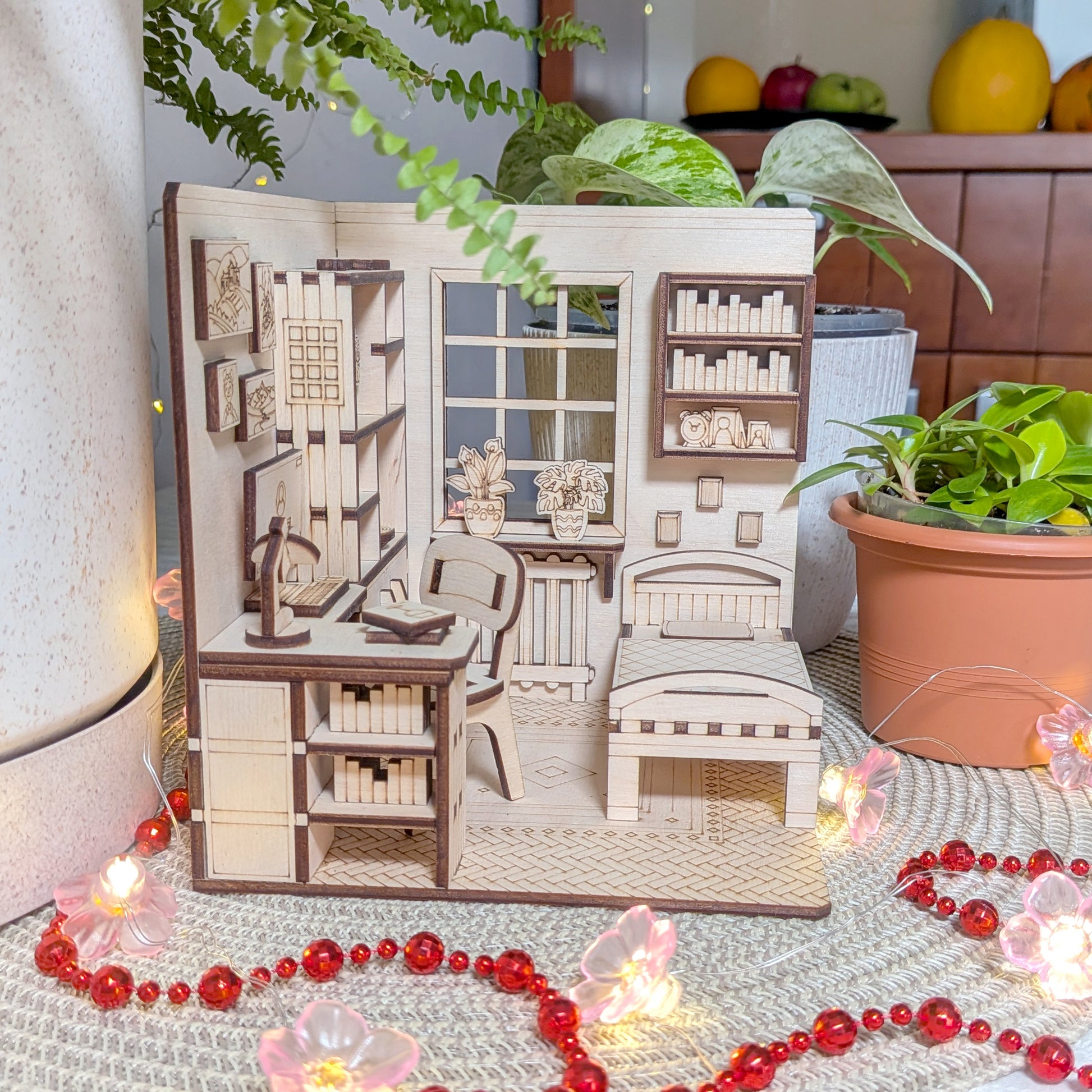 Miniature wooden laser cut model of a room with furniture and decor, surrounded by plants and decorative items.