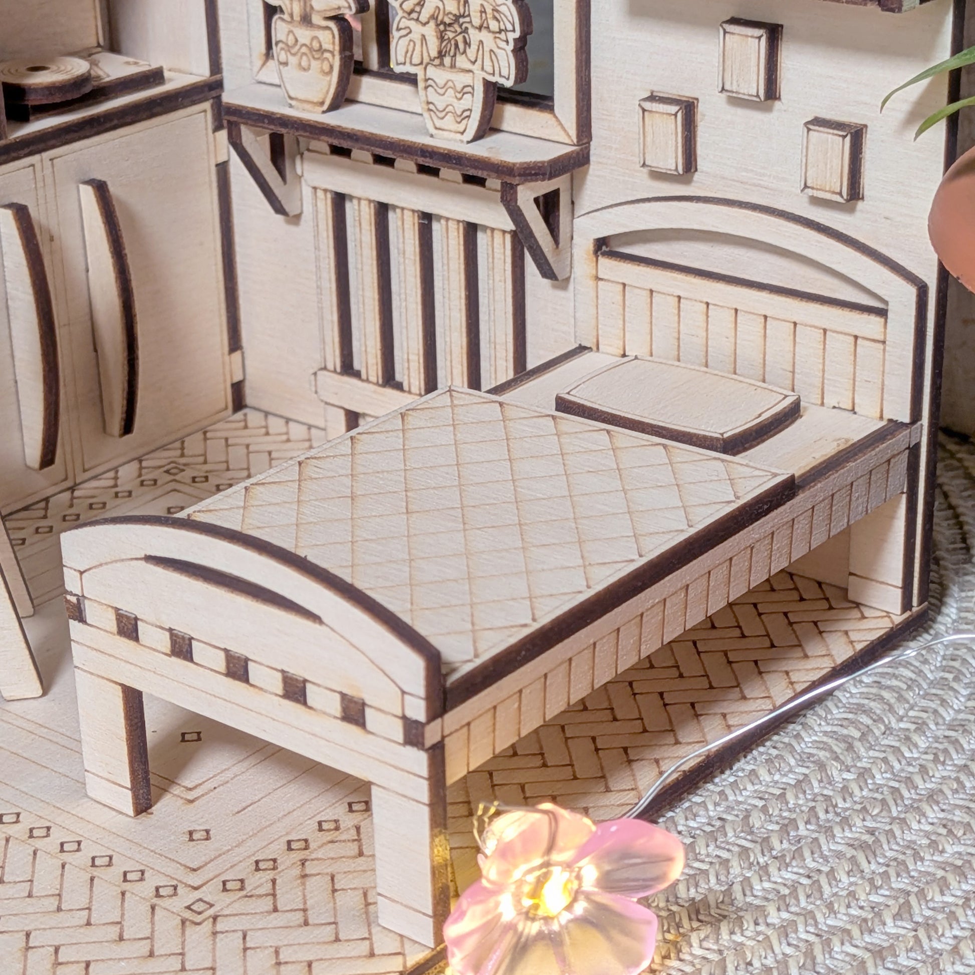 Small wooden miniature bed with etched quilt texture and pillow inside a cozy dollhouse bedroom scene. The bed is next to a window with tiny potted plants on a shelf and a radiator beneath