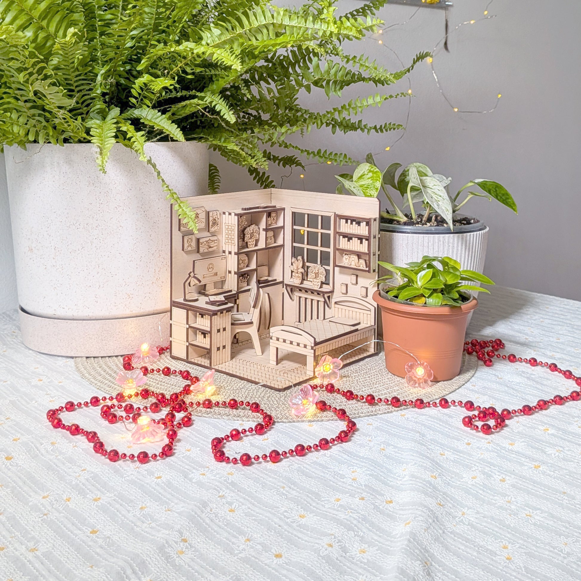 Wooden laser cut model of a room with furniture on a table surrounded by potted plants.