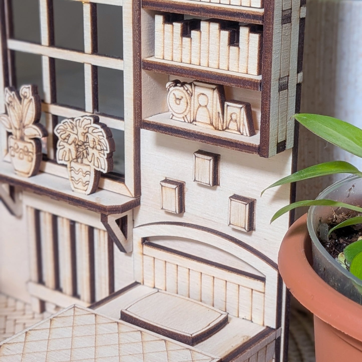 Close-up of a laser cut wooden miniature bedroom scene with a small bed under wall shelves. Tiny books, framed pictures, and potted plants sit beside a window, all etched and layered from light wood