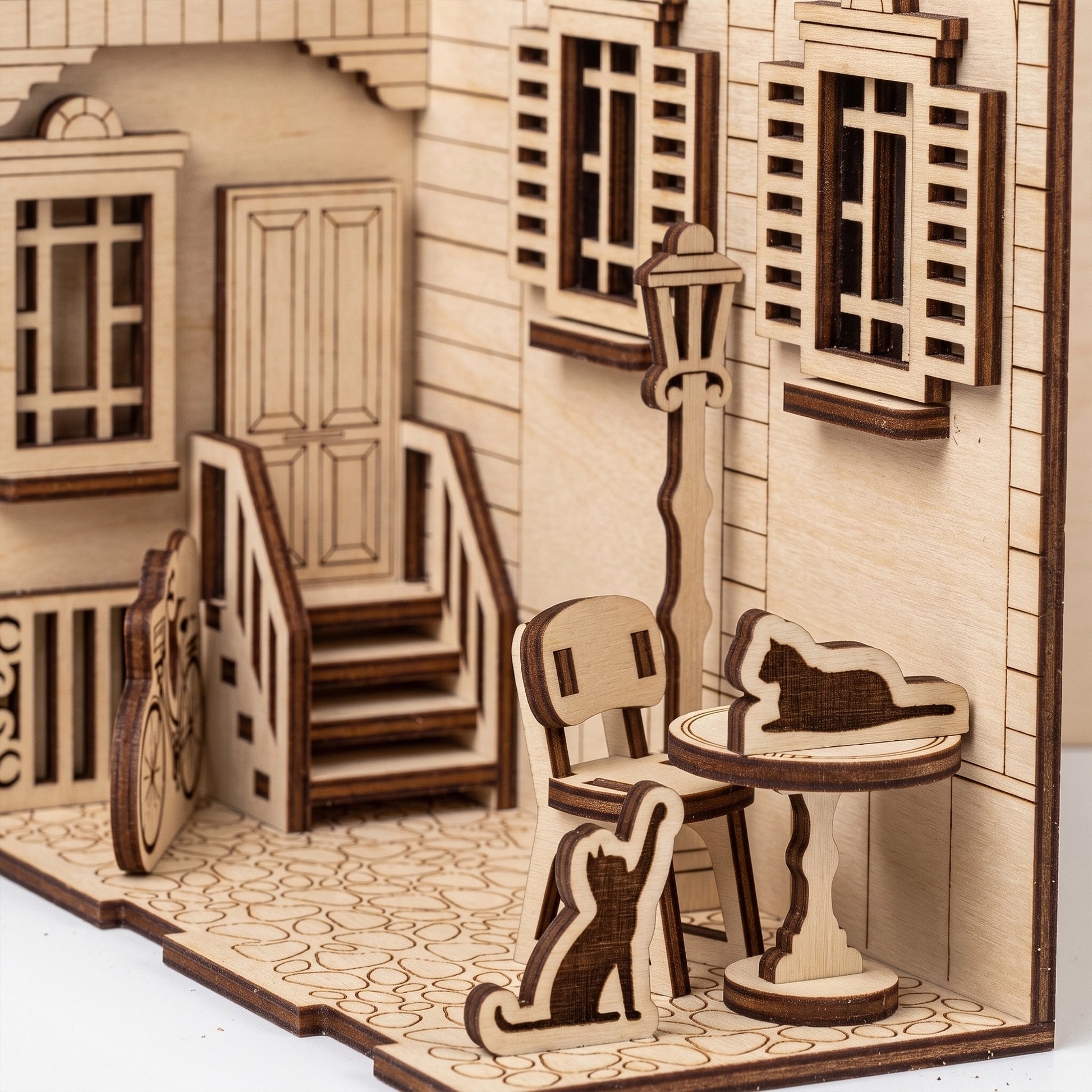 Close-up detail of a laser cut plywood French yard book nook, showing miniature furniture, cat figures, and lamp post