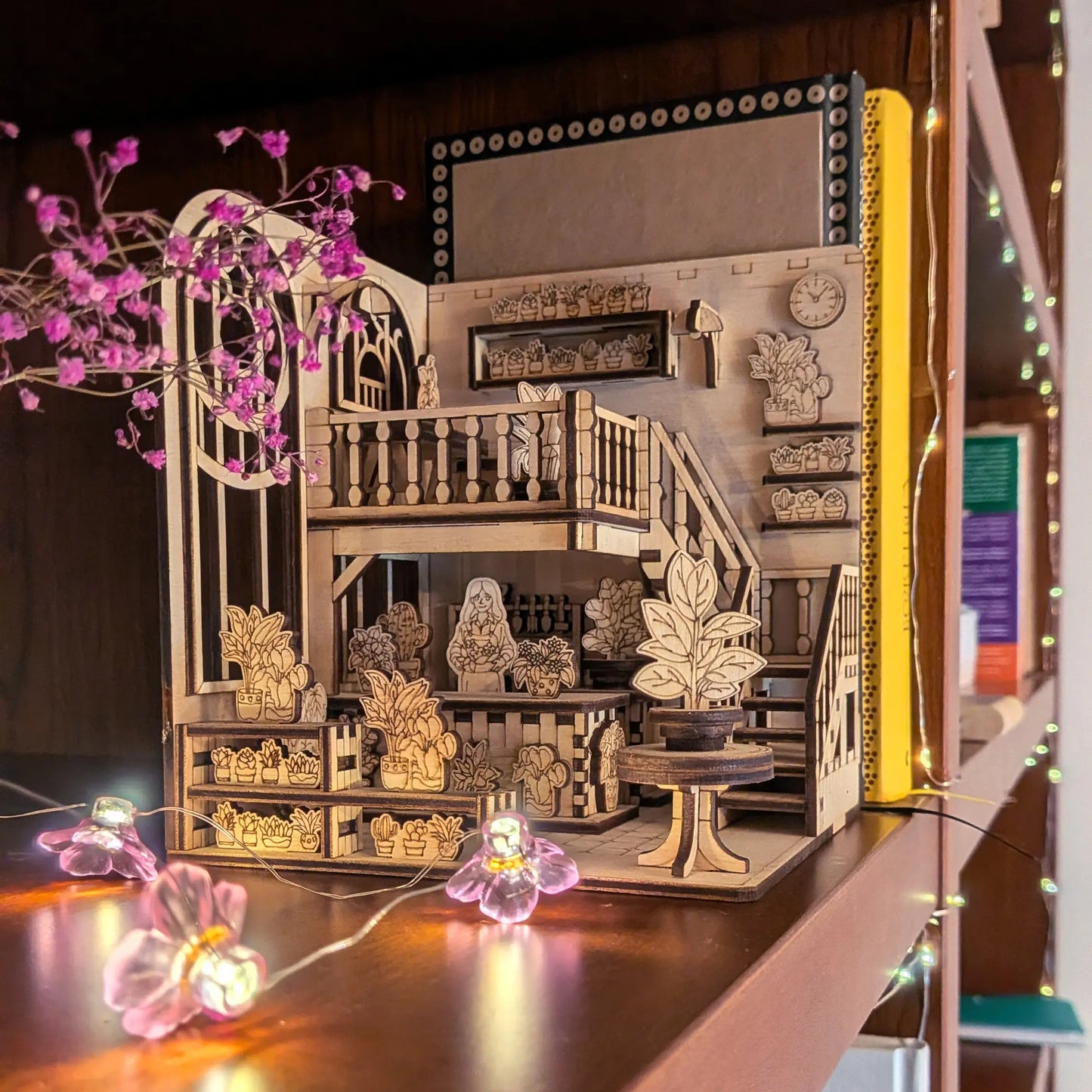 Wooden laser cut Book nook greenhouse dollhouse model with intricate details on a decorative surface with plants.