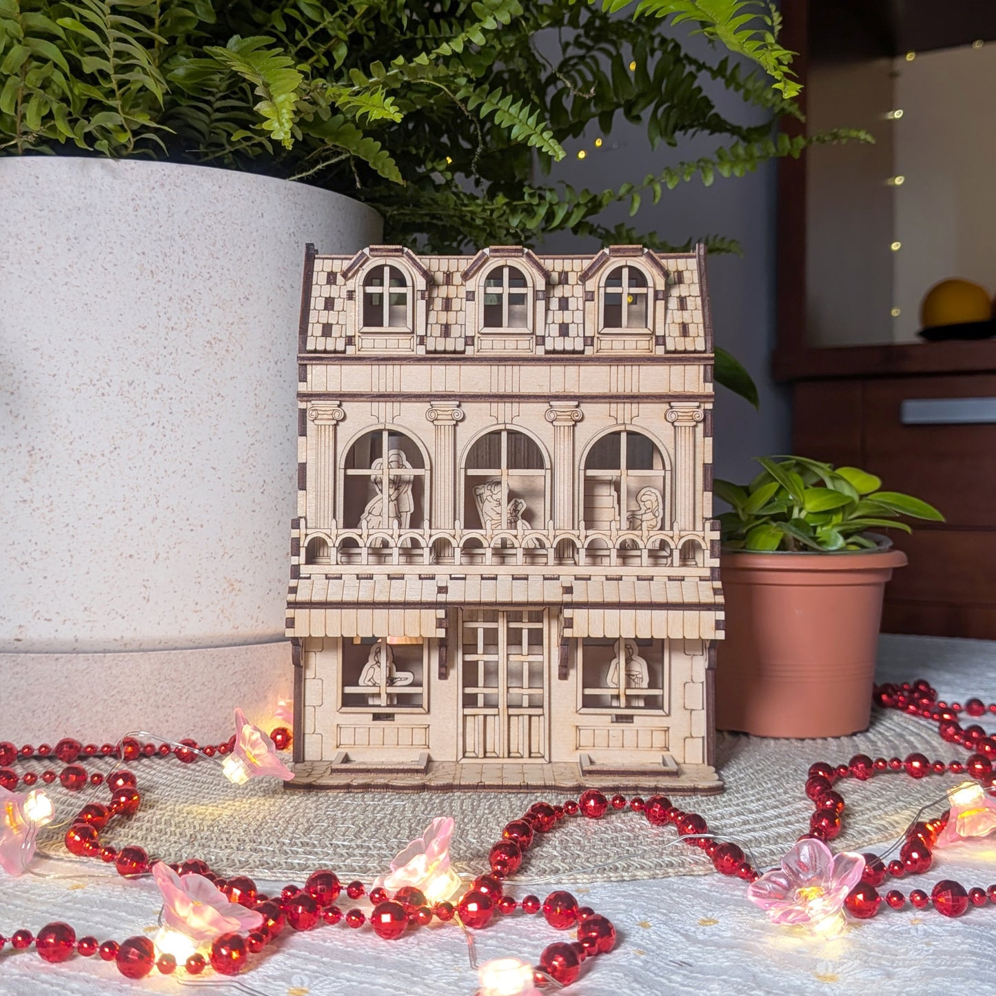 Laser cut townhouse book nook displayed on a white mat with plants and string lights, showing multiple floors and figures inside.