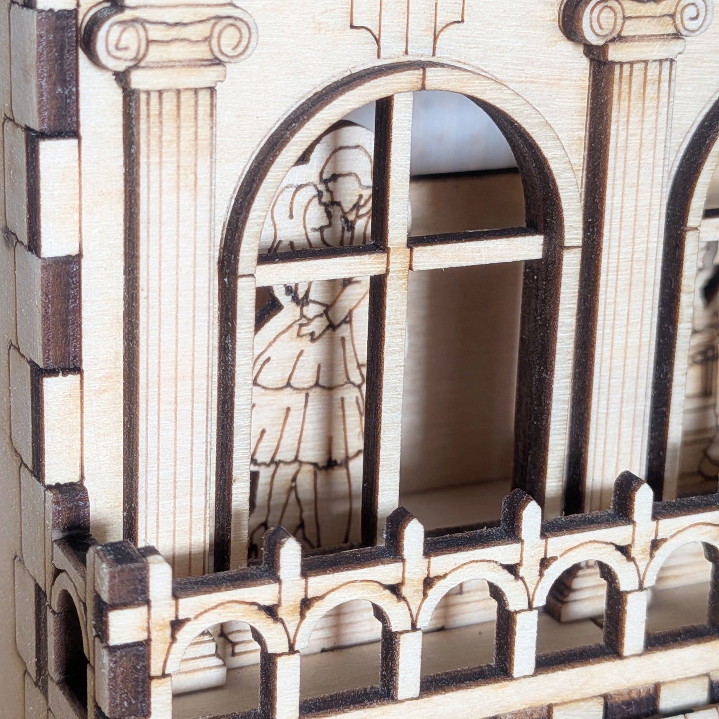 Close-up of a laser cut wooden book nook window showing two small figures inside, framed by engraved columns and arched detailing