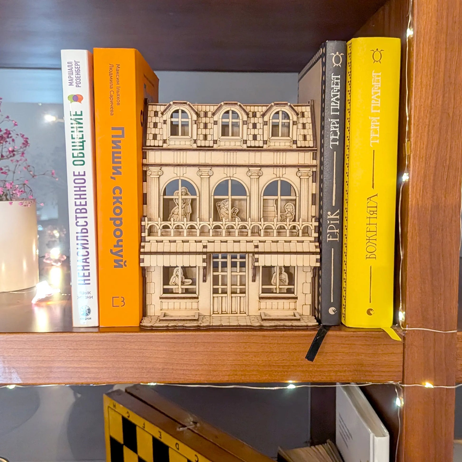 Laser cut plywood book nook house model styled on a bookshelf between colorful books with glowing warm fairy lights