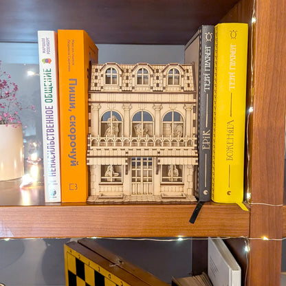 Laser cut plywood book nook house model styled on a bookshelf between colorful books with glowing warm fairy lights