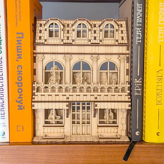 Laser cut wooden book nook shaped like a multi-story townhouse with arched windows and detailed characters inside, placed between books on a shelf.