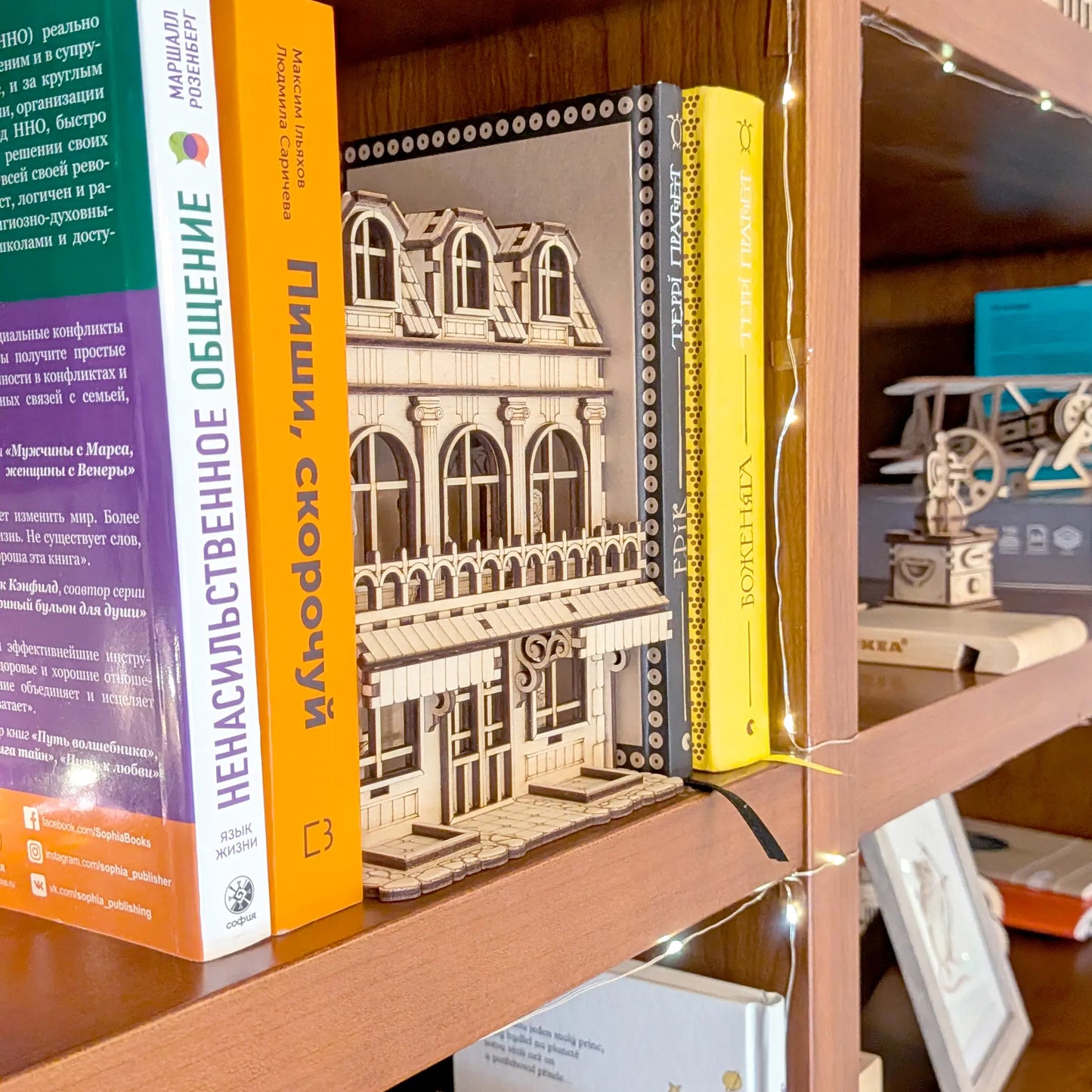 Side angle view of a laser cut plywood house book nook tucked between books on a bookshelf with string fairy lights