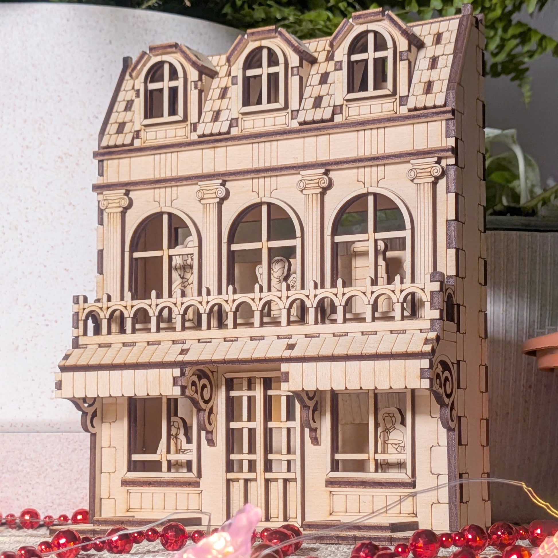Laser cut wooden book nook house displayed with soft lighting and potted plants, showing detailed façade and engraved characters inside the windows