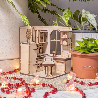 A close view of a laser cut miniature kitchen scene featuring a refrigerator, stove, countertop sink, hanging cooking pans, shelves with tiny dishes, and a round table with a chair. Smooth light wood with engraved details.