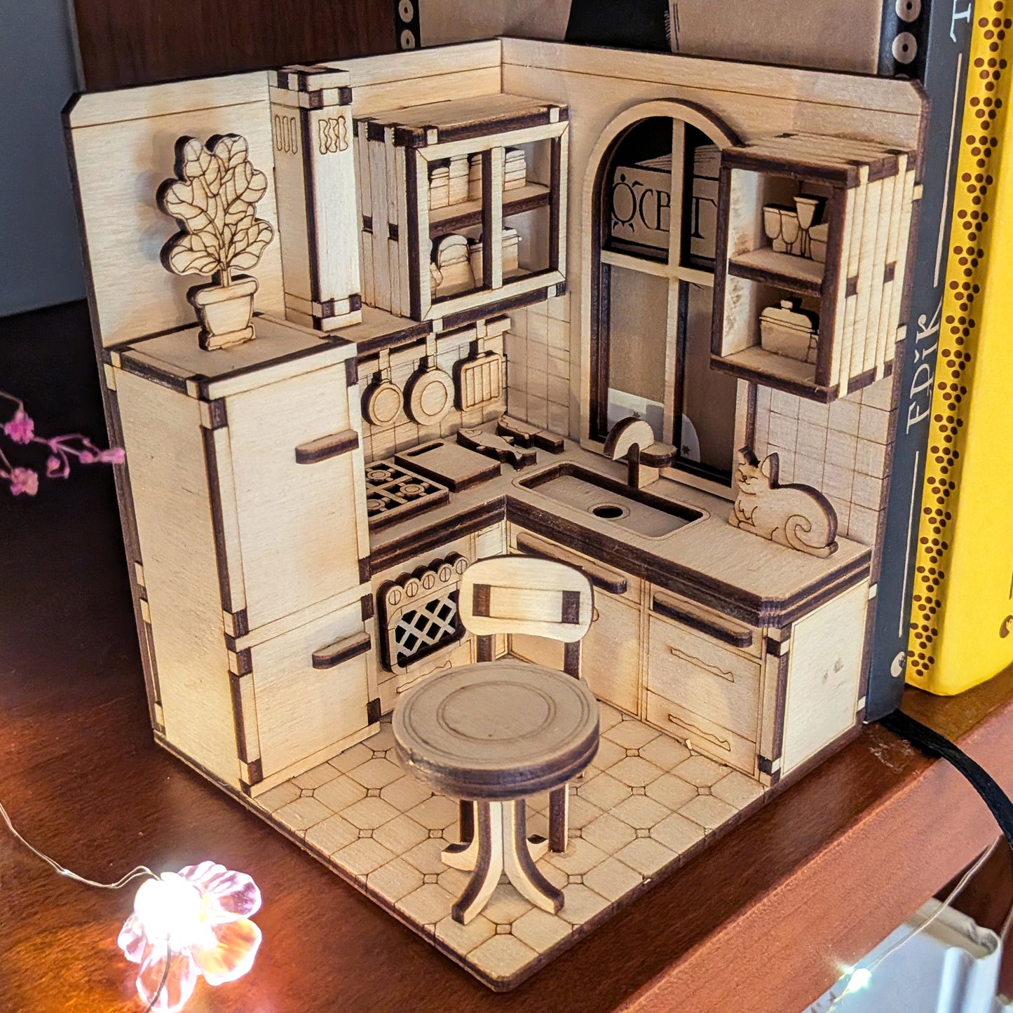Laser cut miniature kitchen book nook scene made of light wood, featuring cabinets, a stove, a chair, shelves, and a small cat figure. Detailed plywood diorama displayed between books.