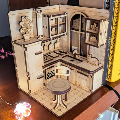 Laser cut miniature kitchen book nook scene made of light wood, featuring cabinets, a stove, a chair, shelves, and a small cat figure. Detailed plywood diorama displayed between books.
