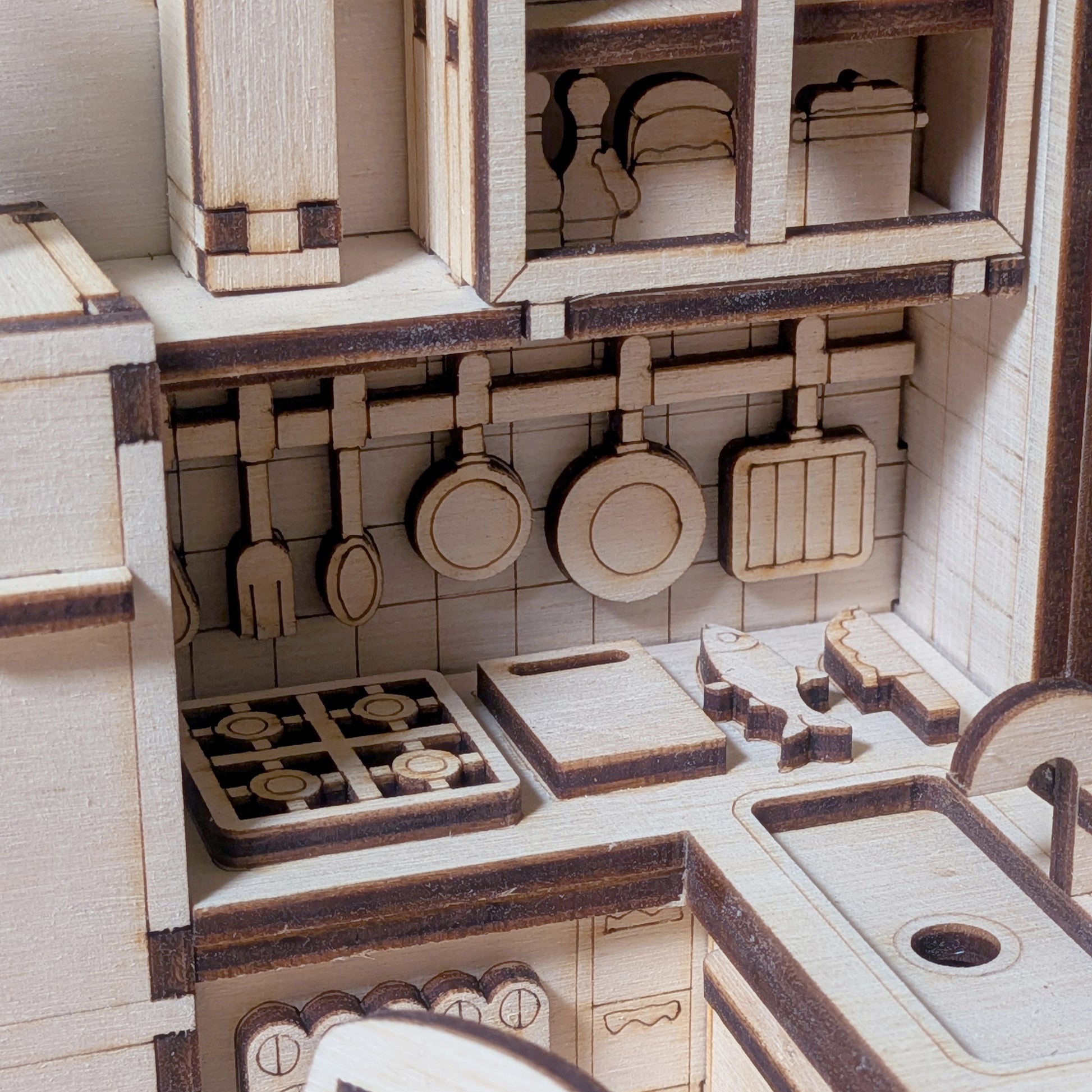 Close-up of laser cut plywood kitchen wall with hanging pans, stove burners, countertop accessories, and engraved cabinet textures