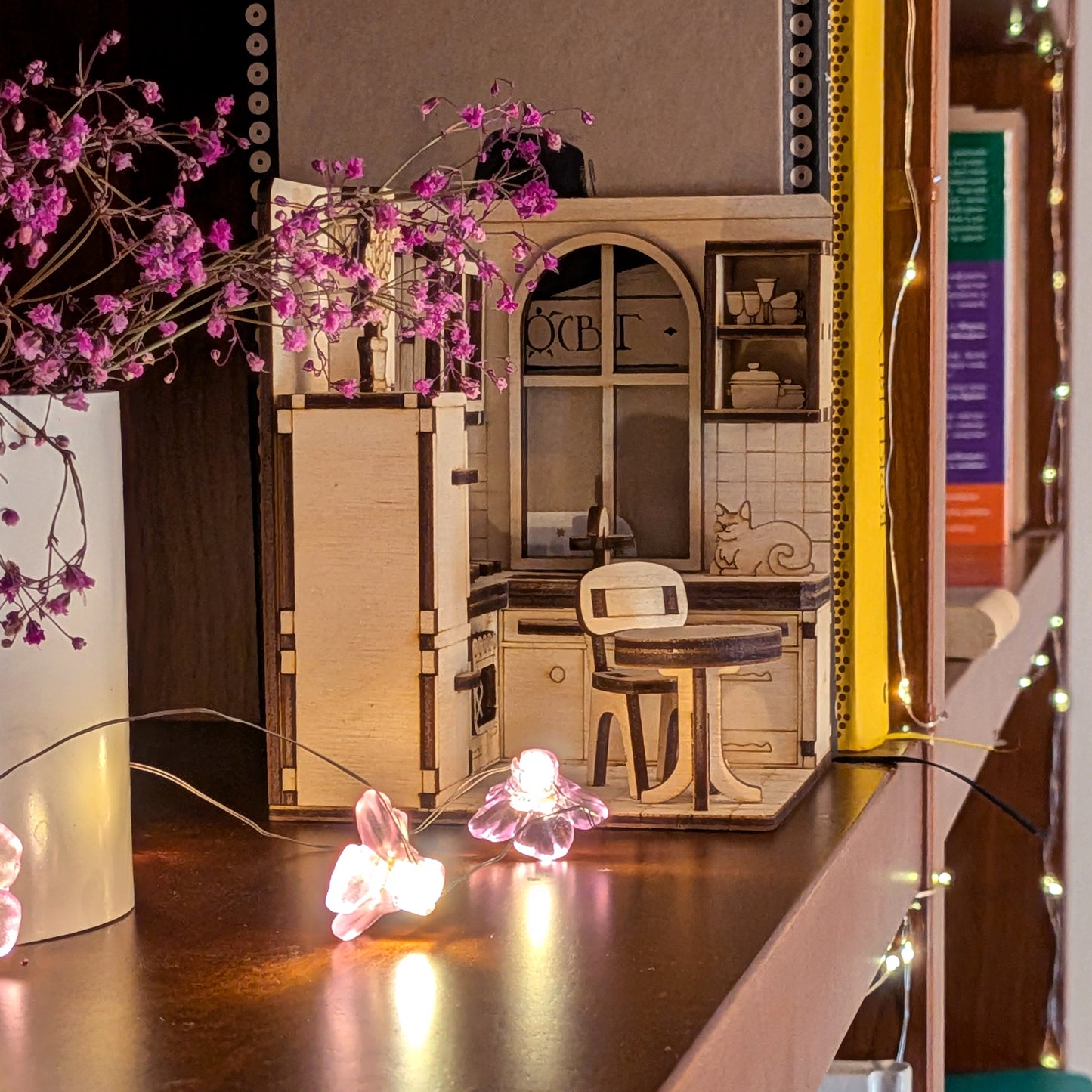Laser cut miniature kitchen book nook scene on a shelf, with small wooden cabinets, a chair, a counter, and a cat detail. Displayed with flower lights.