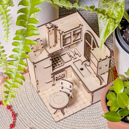 Top-down view of a detailed miniature kitchen diorama made from plywood. Visible are the stove burners, cutting boards, sink, countertop edges, wall utensils, and a round table with a chair below.