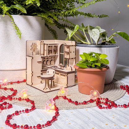 A small wooden kitchen diorama placed among potted plants and decorative lights. The laser cut miniature includes cabinets, a round table, a chair, and a cat sculpture near the sink, creating a cozy indoor scene