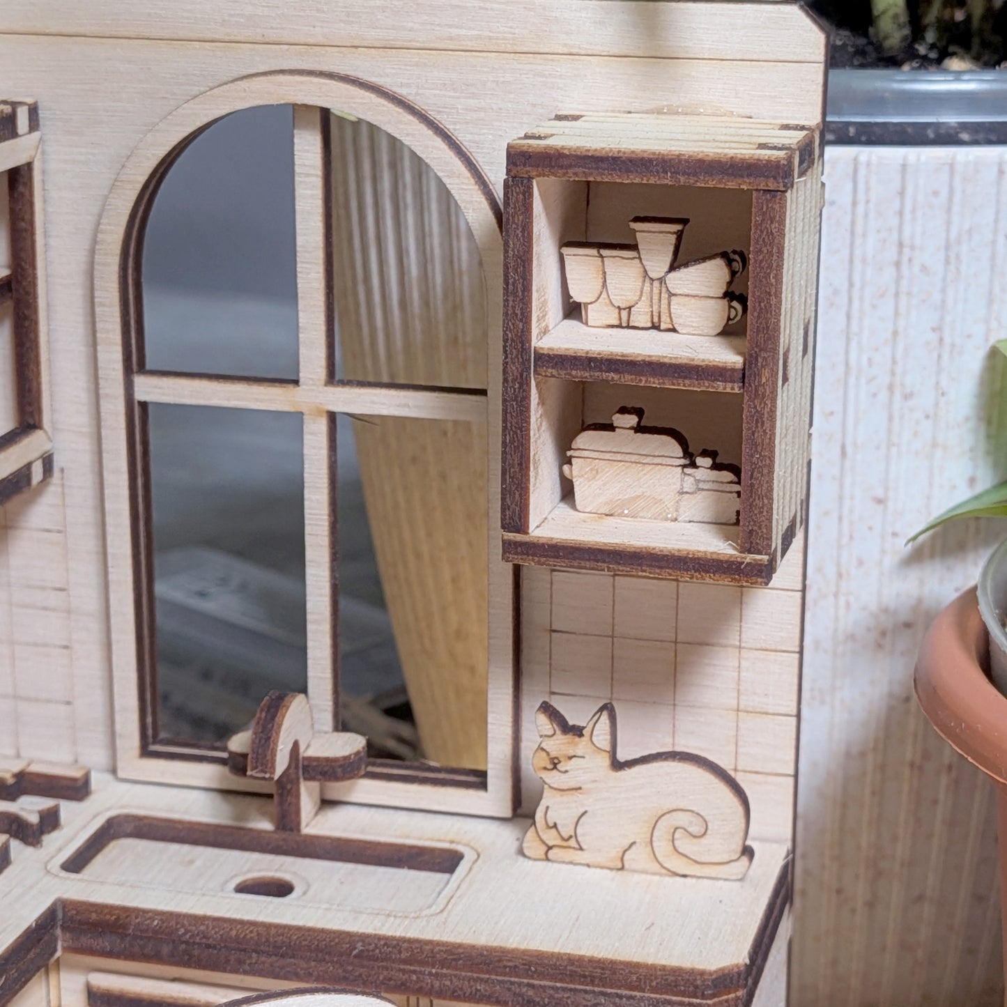 Laser cut plywood kitchen detail with a window, countertop sink, open shelves of dishes, and a small wooden cat figure sitting near the counter.