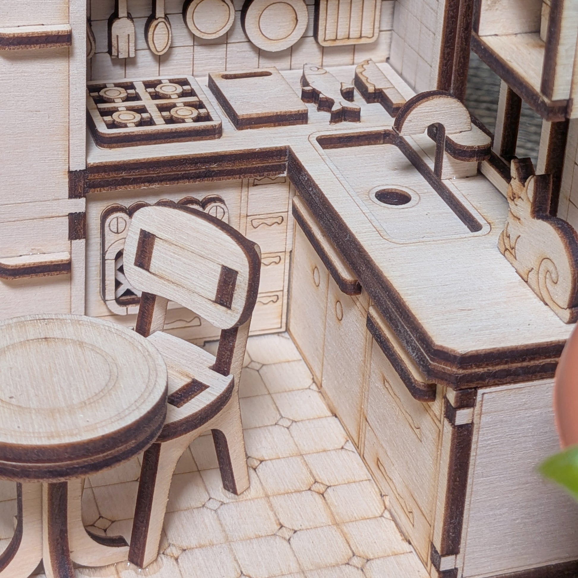 Close-up of a laser cut miniature kitchen diorama made from plywood, showing cabinets, a stove, utensils on the wall, and a small table with a chair.
