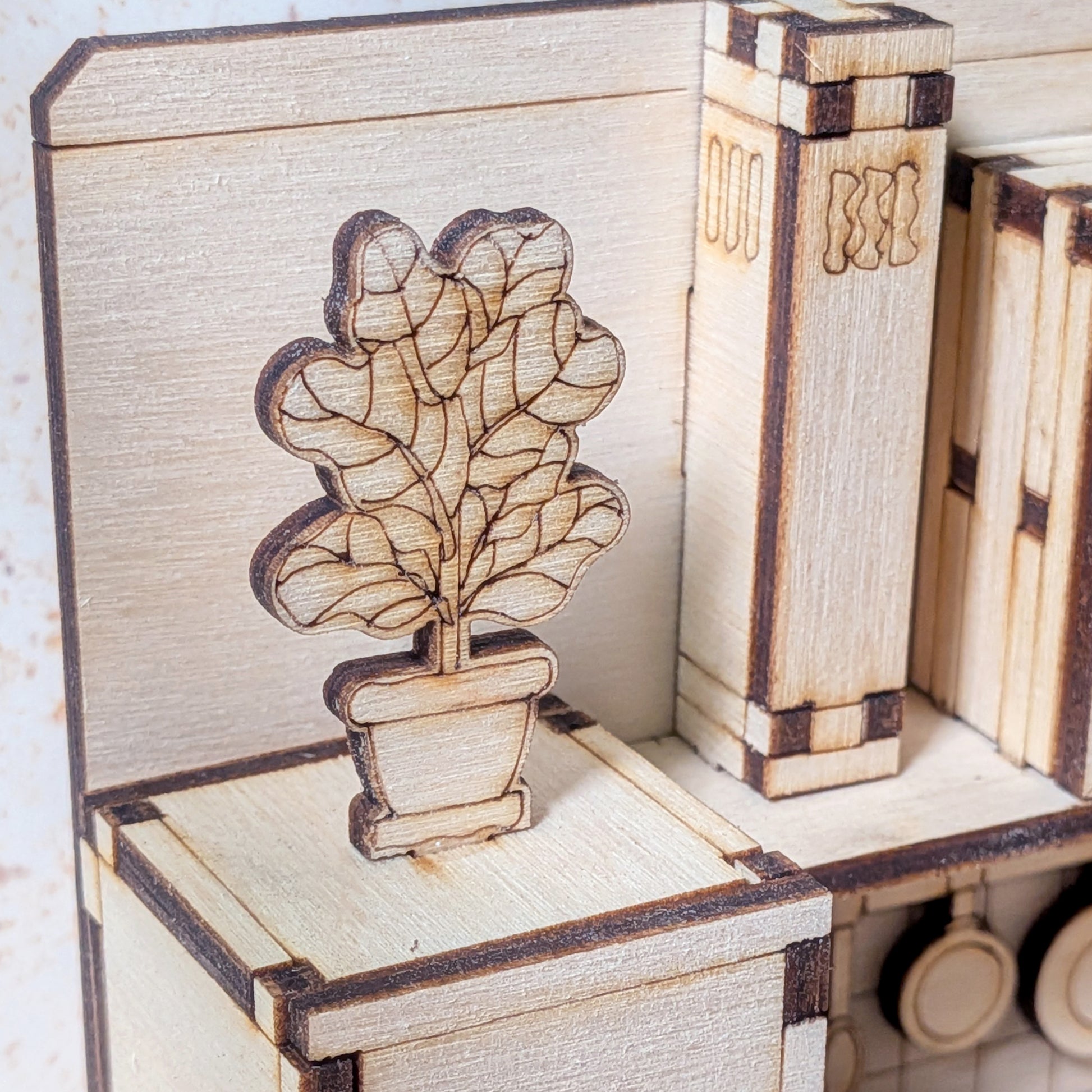 A close-up of a small laser cut wooden plant in a pot, sitting on top of a miniature kitchen cabinet. The plant and pot are made from light plywood with engraved leaf details and soft burn marks from the laser cutting process.