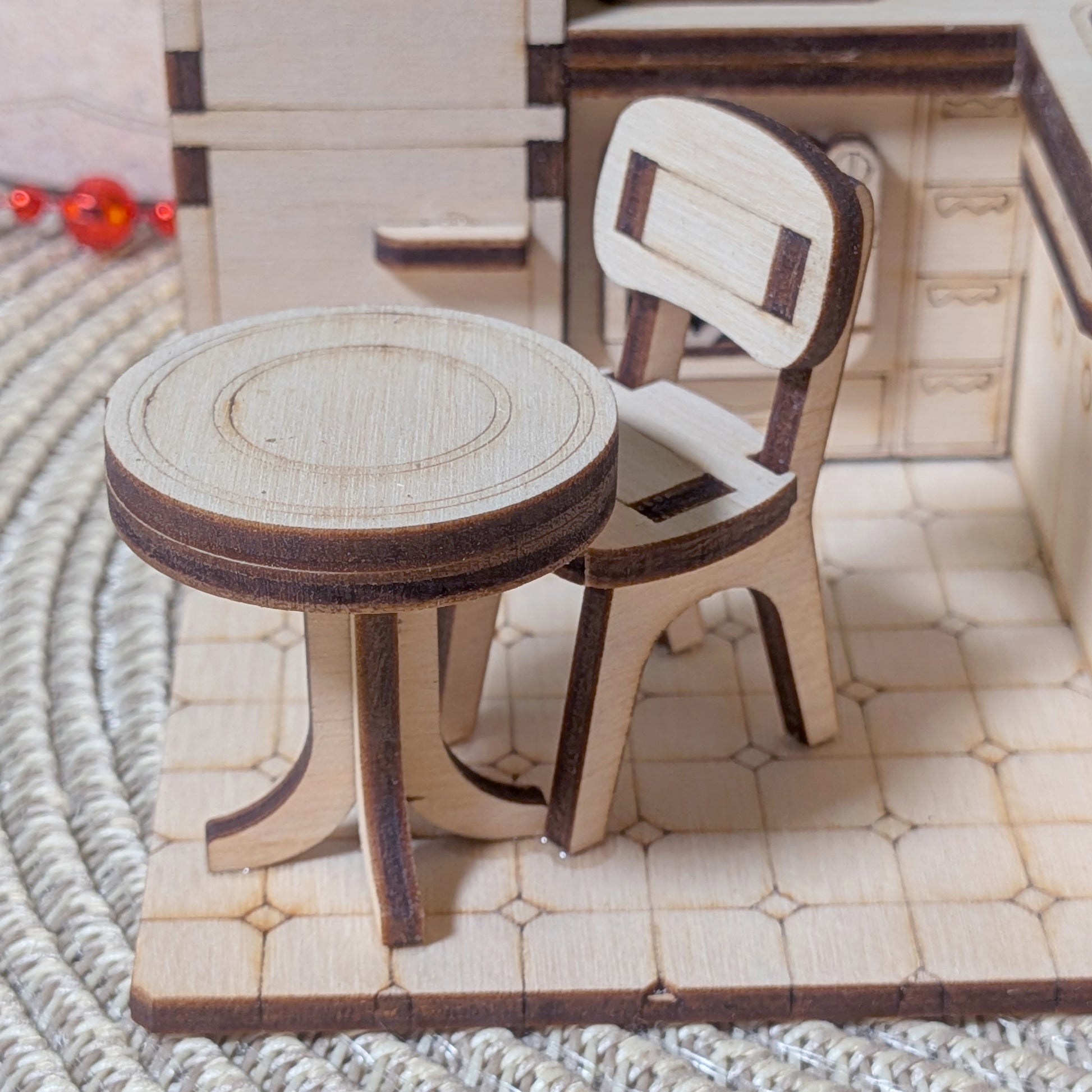 Laser cut miniature wooden table and chair placed on a tiled floor inside a tiny kitchen model. The furniture is made from layered plywood pieces with visible dark laser edges and engraved circular surface details