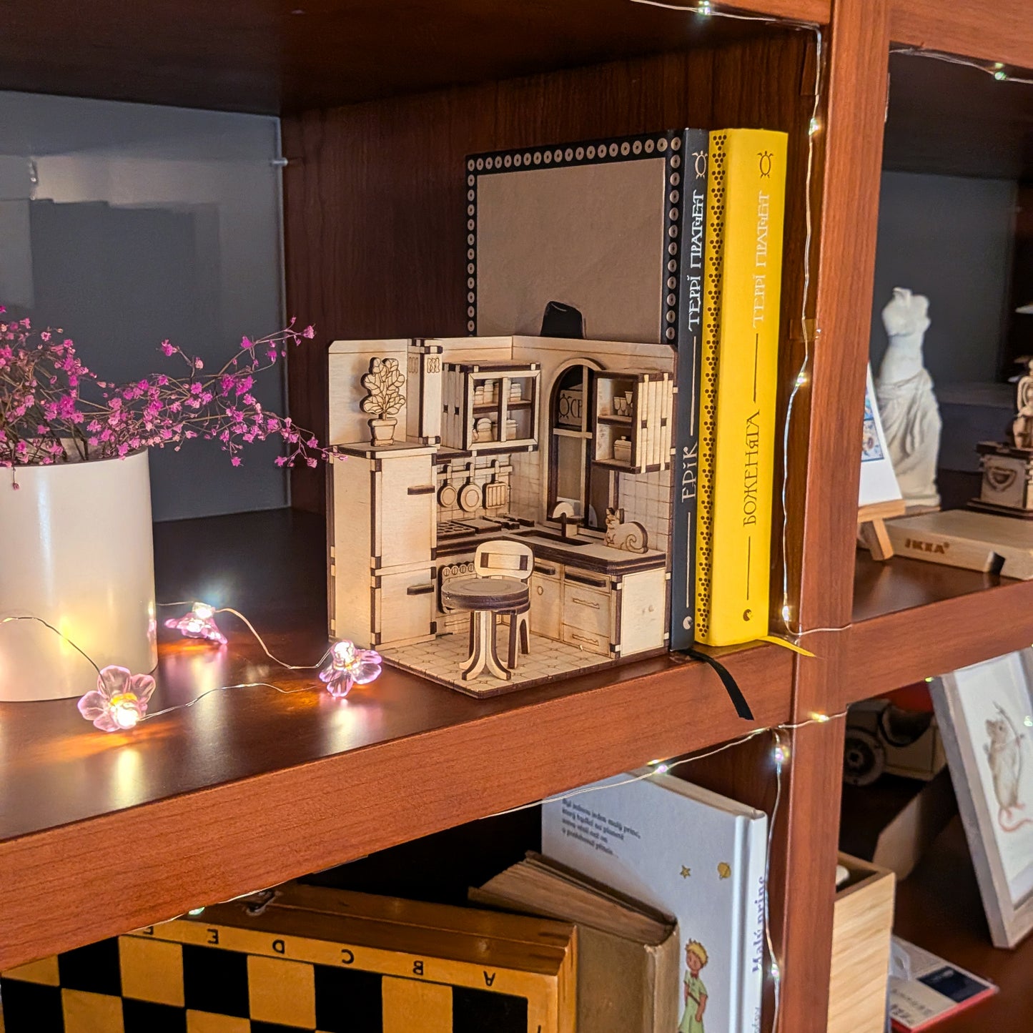 A miniature laser cut wooden kitchen diorama displayed on a bookshelf. The scene includes cabinets, a stove, table, chair, and a small cat figure on the counter. Warm string lights and books surround it, creating a cozy atmosphere.
