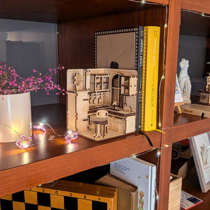 A miniature laser cut wooden kitchen diorama displayed on a bookshelf. The scene includes cabinets, a stove, table, chair, and a small cat figure on the counter. Warm string lights and books surround it, creating a cozy atmosphere.