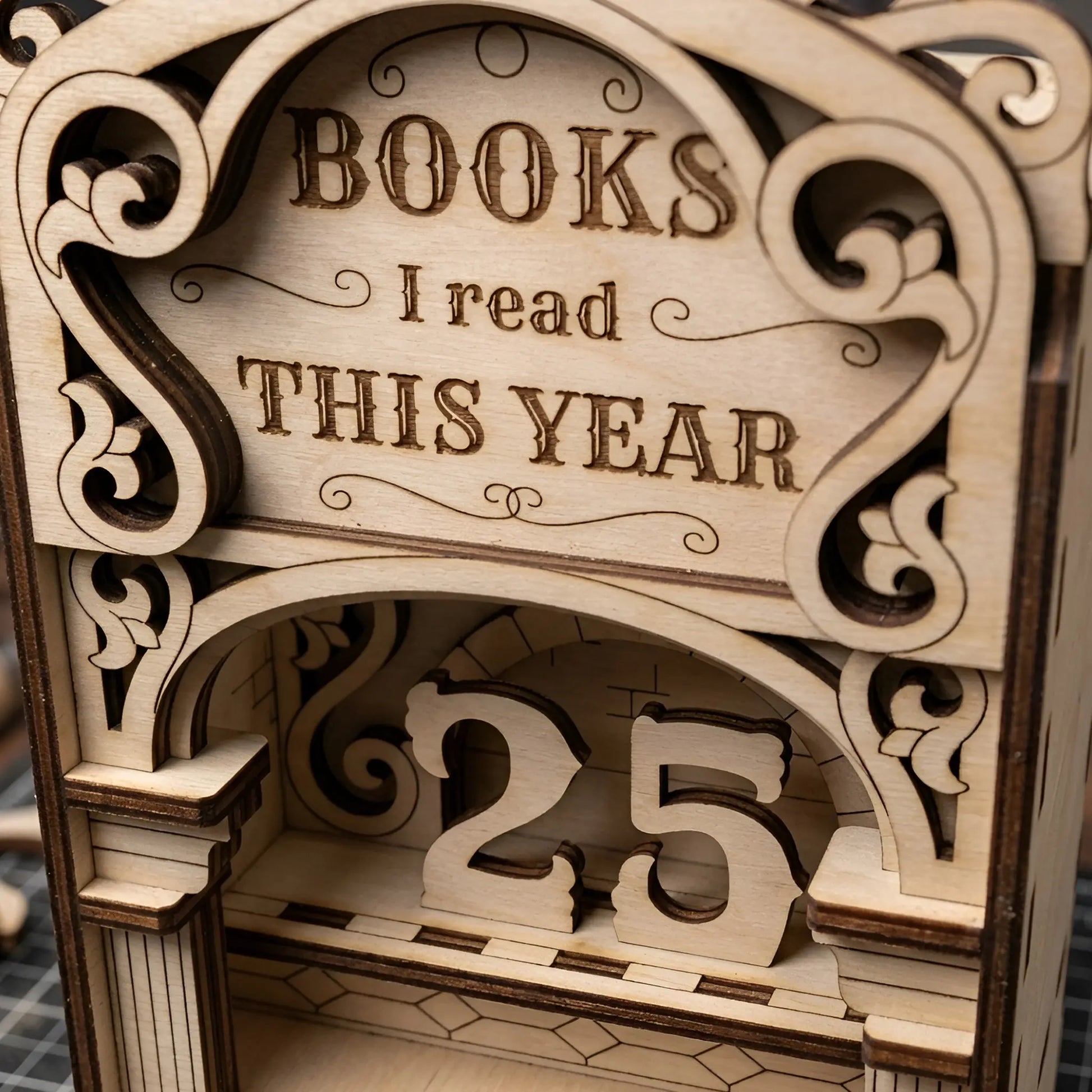 Close-up of a laser cut wooden book reading tracker with engraved lettering, decorative scrollwork, arch design, and removable number pieces showing 25 books read this year.