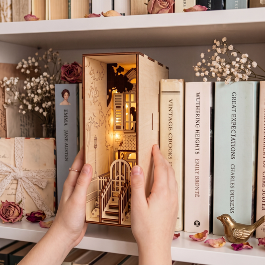 Hands placing a lit laser-cut birch plywood romantic house book nook on a shelf between classic novels.