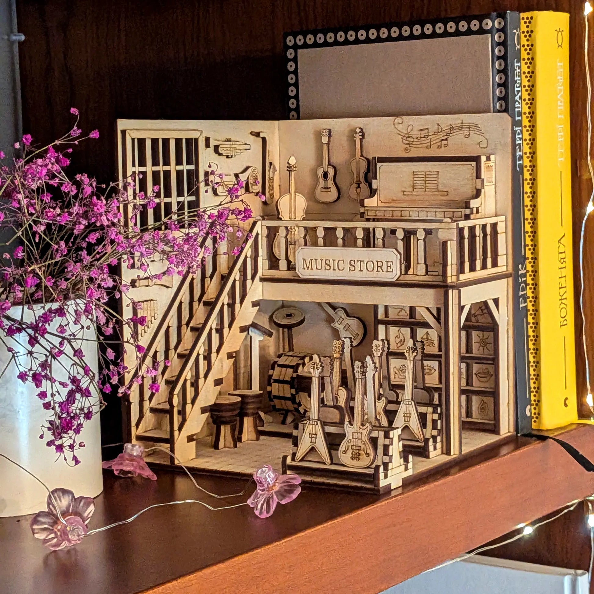 Laser cut plywood music store book nook on a dark wood shelf next to violet flowers and colorful book spines.