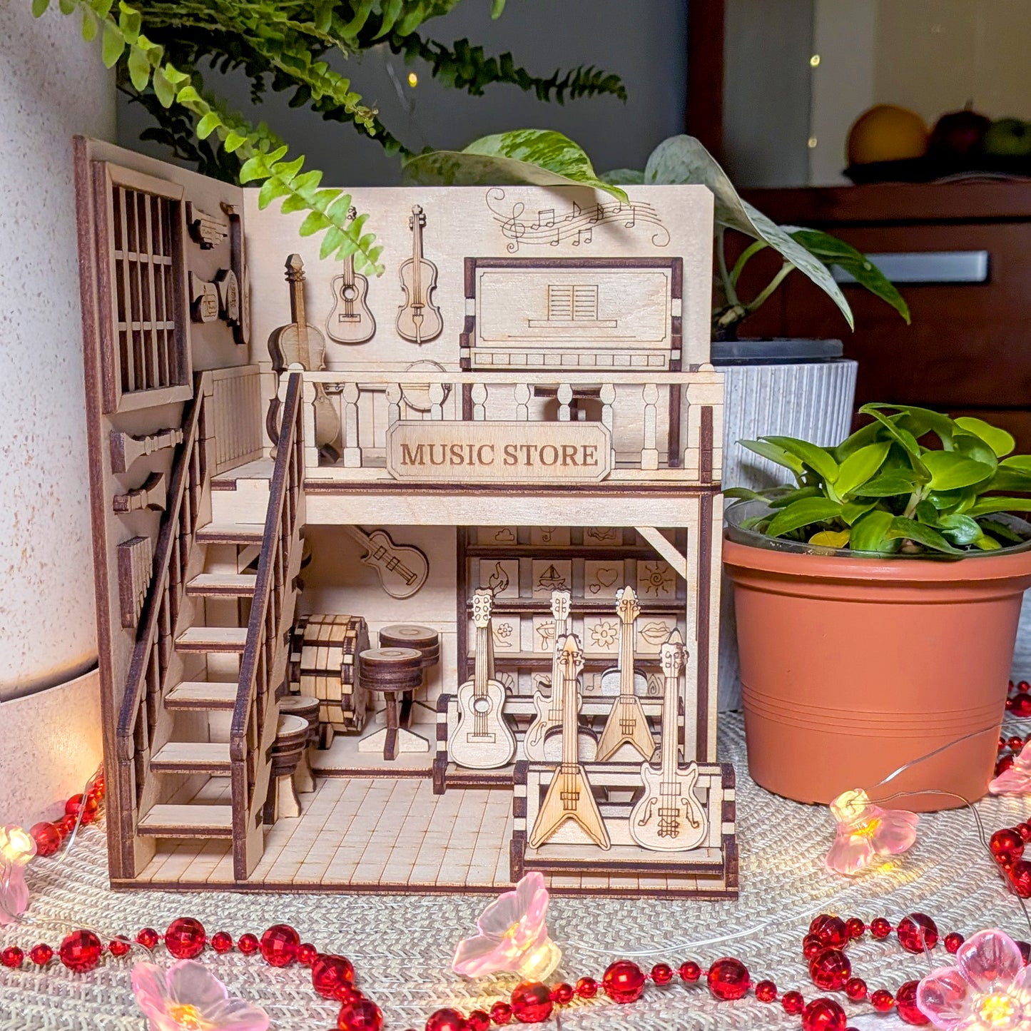 Laser cut wooden book nook diorama of a miniature music store, featuring guitars, drums and instruments, displayed on a table with plants and decorative string lights