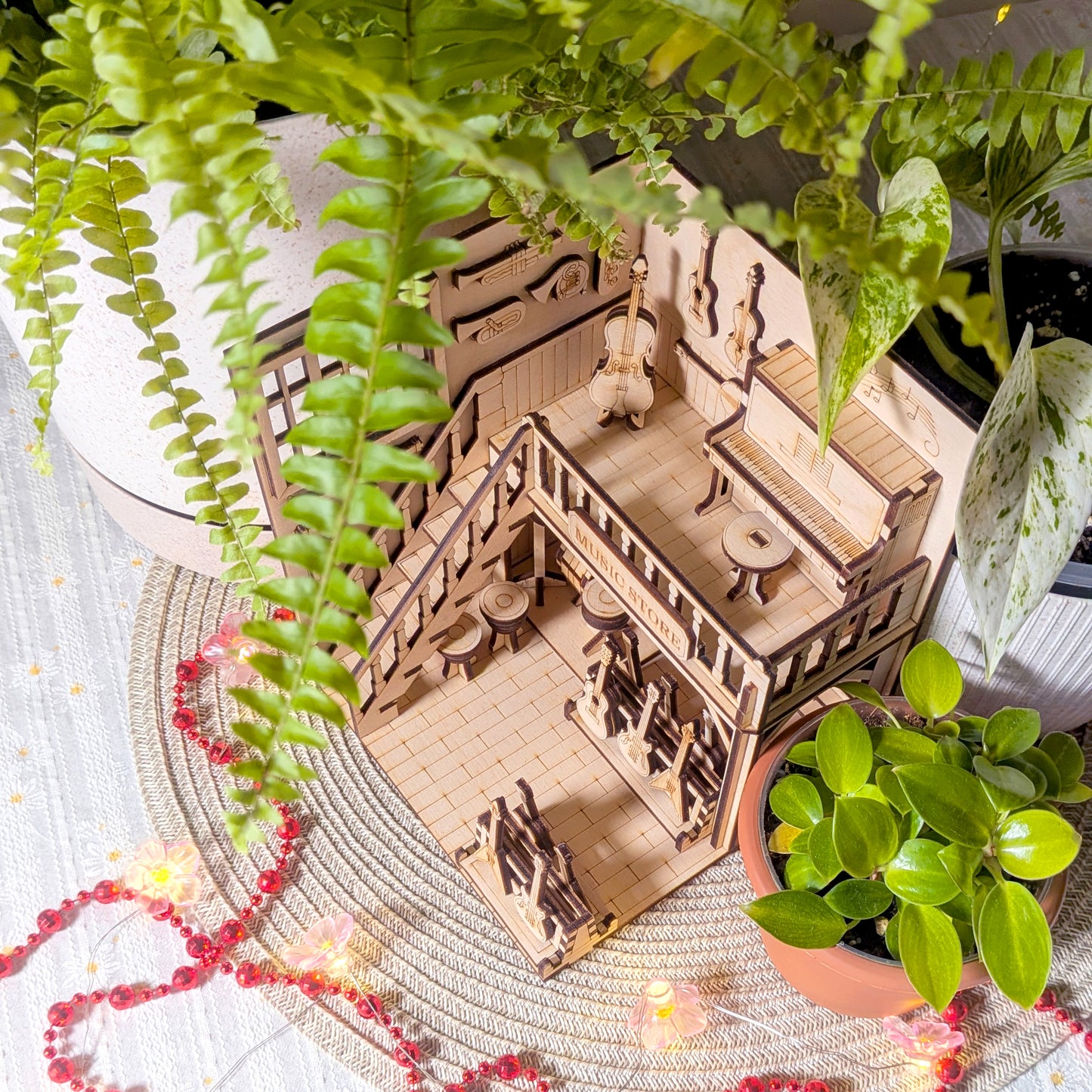 Top angled view of a laser cut wooden music store diorama showing the upper piano area, guitars and interior details surrounded by green plants and string lights