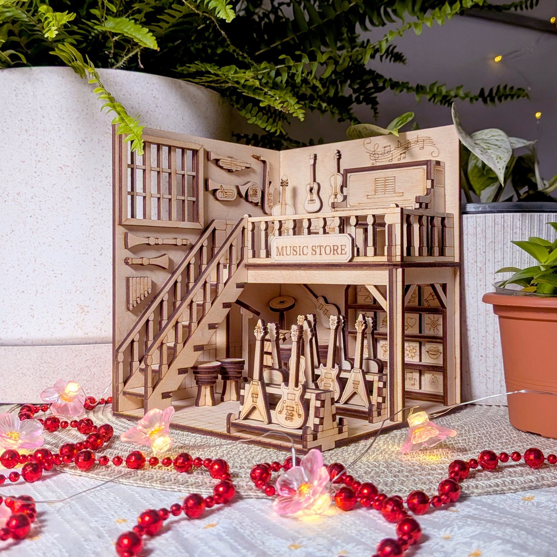 Front view of a laser cut music store book nook with detailed stairs, guitars and drums, placed beside potted plants and red beaded lights