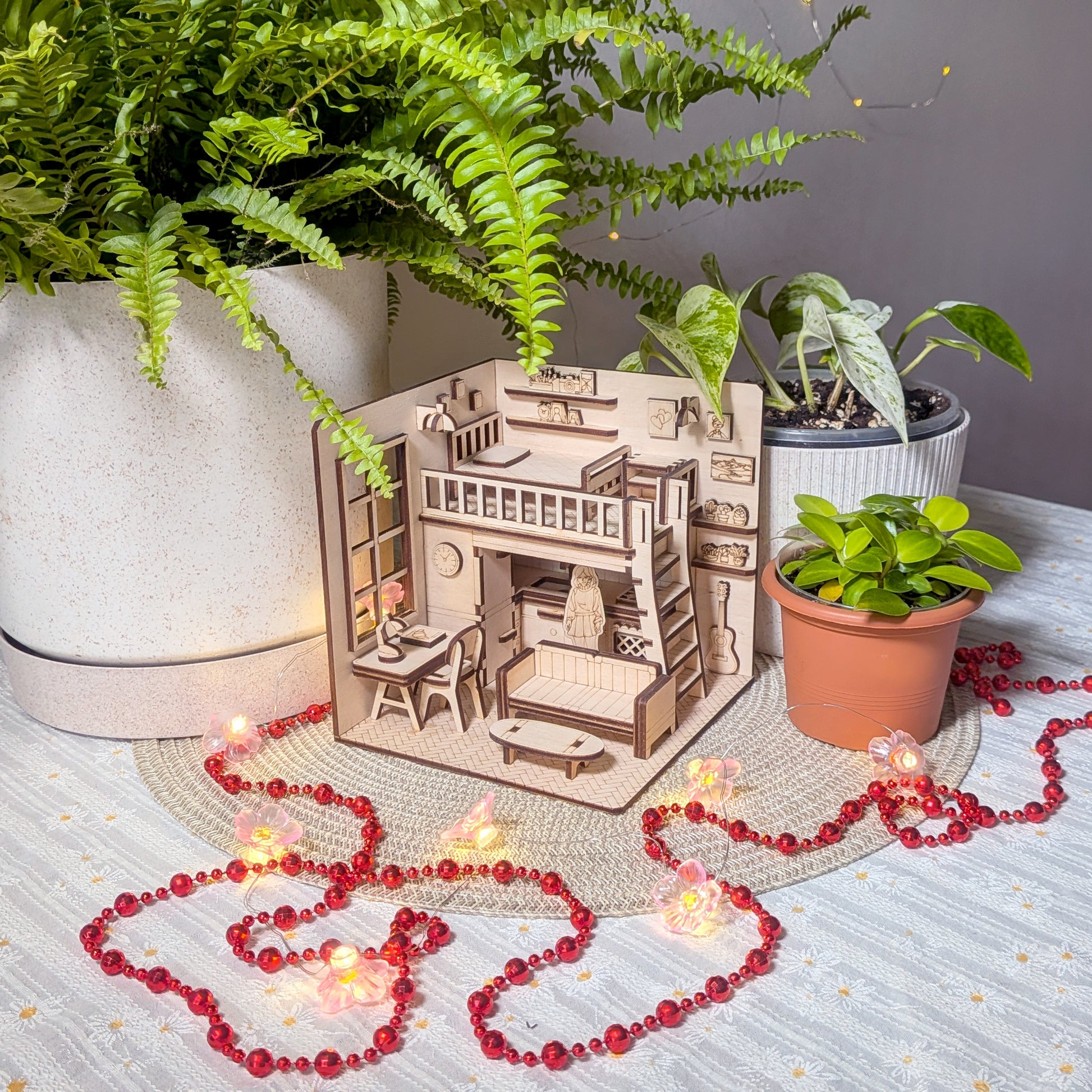 Miniature wooden dollhouse room diorama with loft bed, desk, sofa and shelves, displayed among plants and fairy lights. Designed as a laser cut book nook template and DIY wooden project