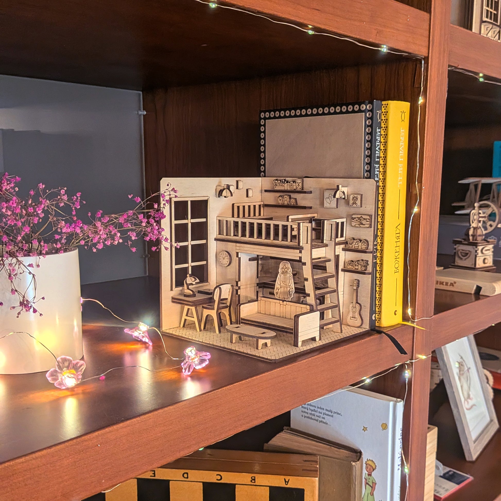 Miniature wooden dollhouse-style book nook displayed on a bookshelf, with loft bed, desk and sofa details. Designed as a laser cut wooden diorama template for DIY assembly
