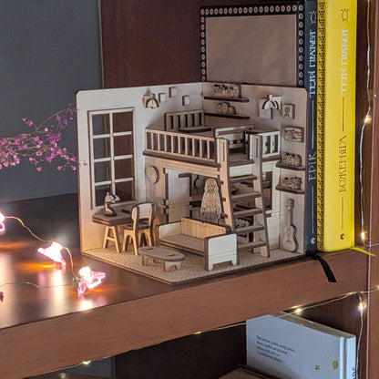 Miniature wooden dollhouse-style book nook displayed on a bookshelf, with loft bed, desk and sofa details. Designed as a laser cut wooden diorama template for DIY assembly