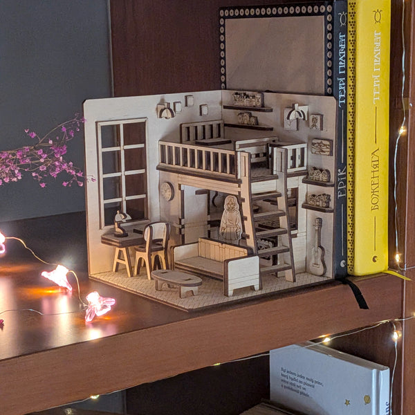 Miniature wooden dollhouse-style book nook displayed on a bookshelf, with loft bed, desk and sofa details. Designed as a laser cut wooden diorama template for DIY assembly