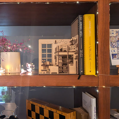 Miniature wooden dollhouse room book nook displayed on a bookshelf next to books and decor. Features loft bed, desk and sofa details. Made as a laser cut wooden diorama template for DIY assembly