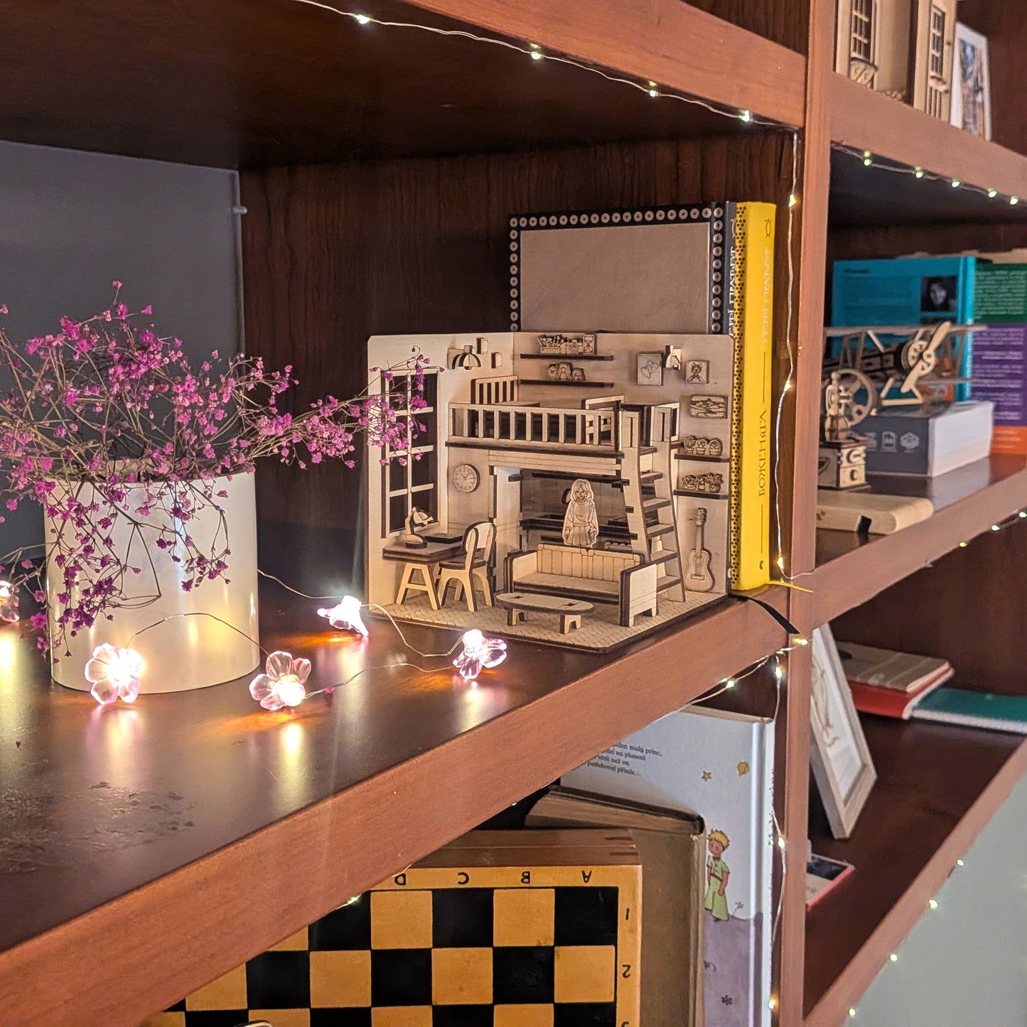Wooden dollhouse-style book nook displayed on a bookshelf, surrounded by fairy lights and decor. The miniature room features a loft bed, furniture and details, made from a laser cut diorama template