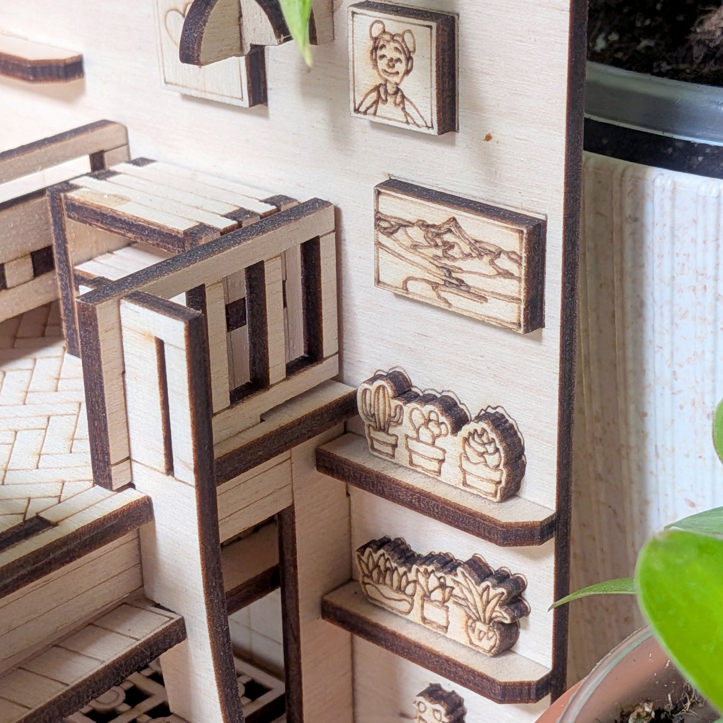 Close-up of miniature wooden wall shelves with engraved potted plants and framed artwork in a dollhouse room. Part of a laser cut wooden diorama and book nook template