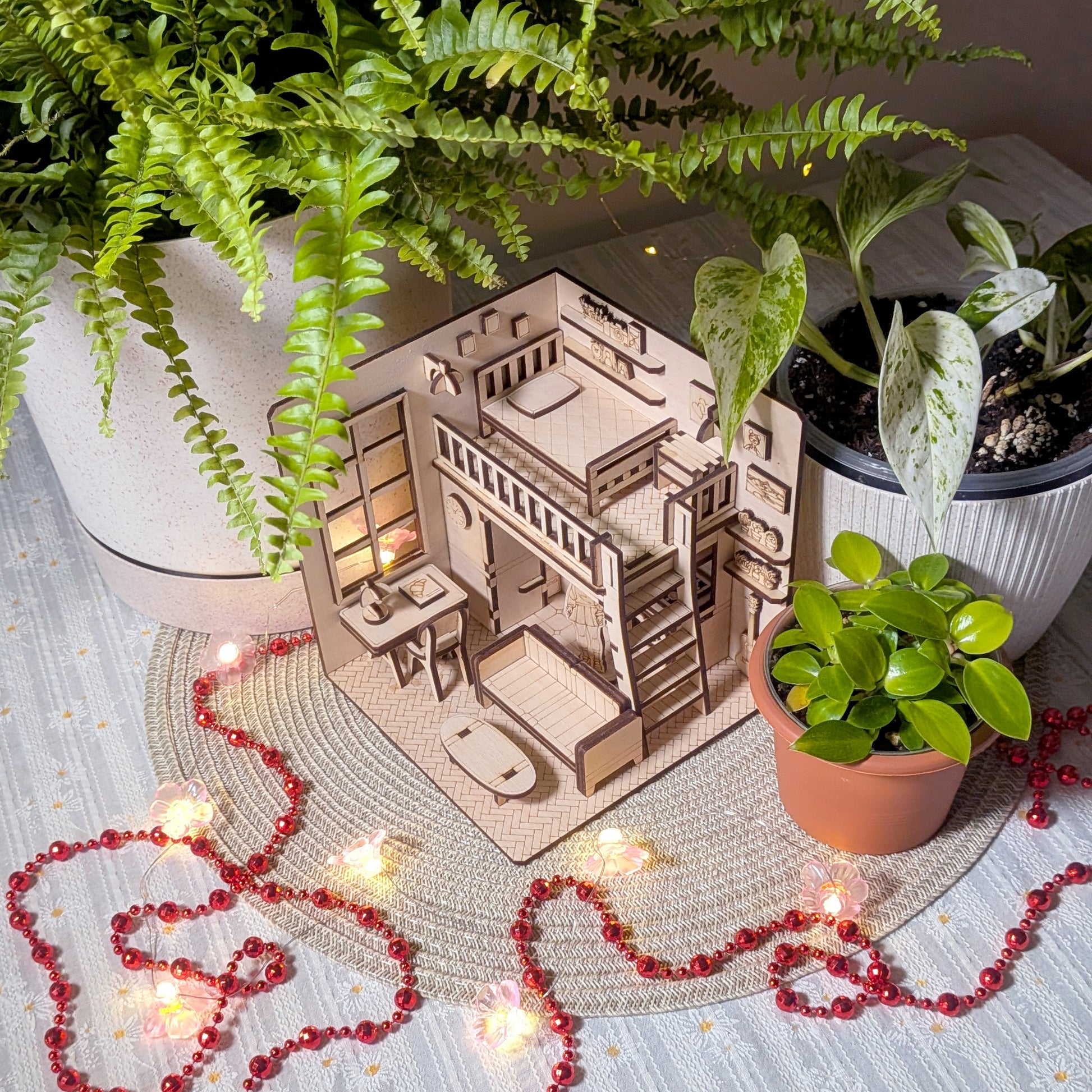 Miniature wooden dollhouse room diorama with loft bed, desk and sofa, displayed among potted plants and fairy lights. Designed as a laser cut book nook template for a DIY wooden project