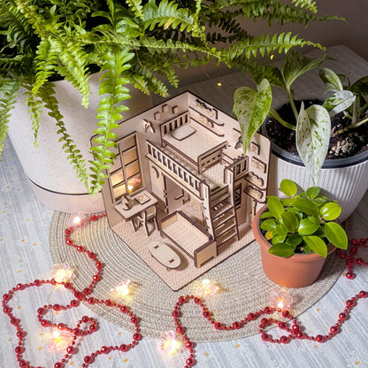 Miniature wooden dollhouse room diorama with loft bed, desk and sofa, displayed among potted plants and fairy lights. Designed as a laser cut book nook template for a DIY wooden project