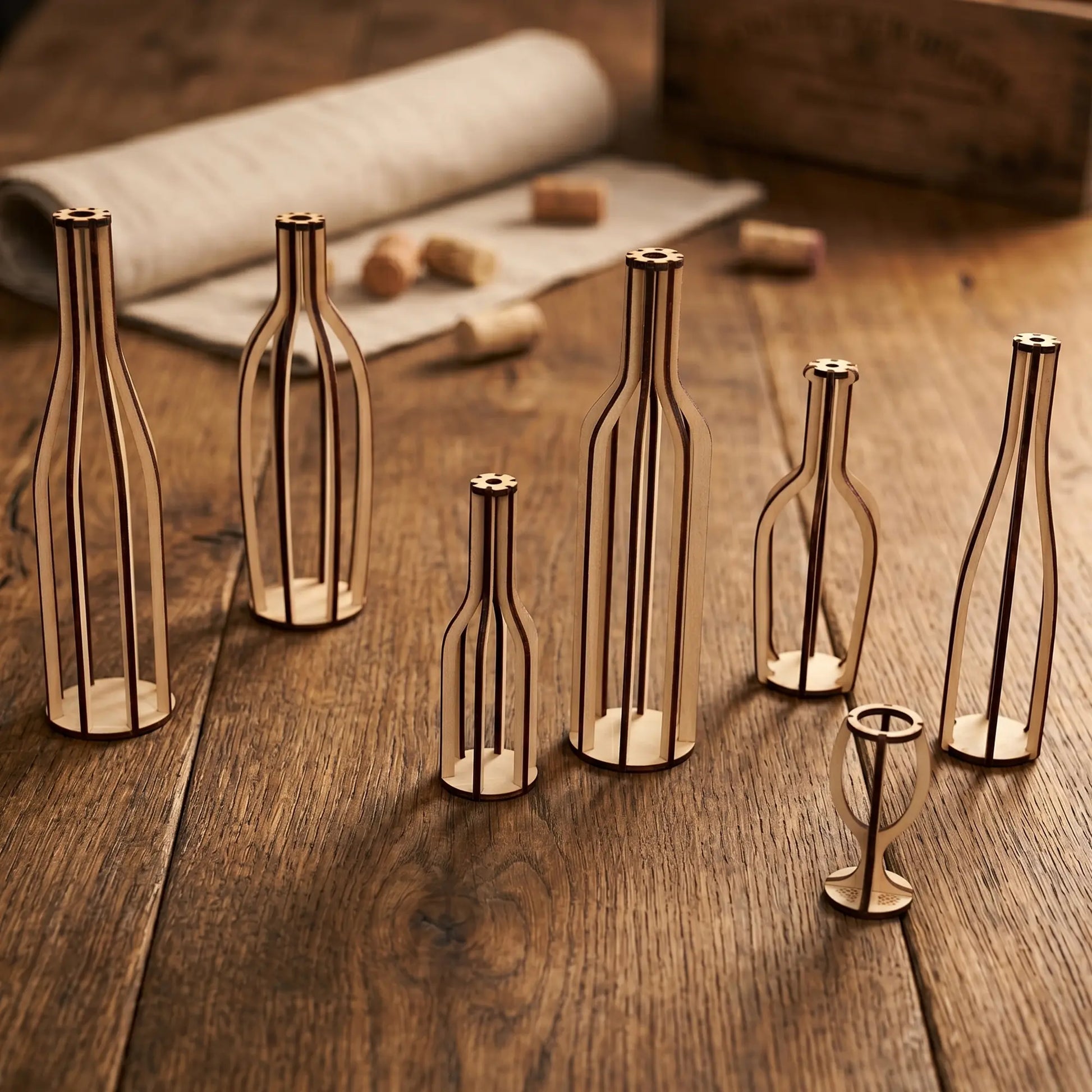 Skeletal laser cut plywood bottle silhouettes in various shapes on a rustic dark wood table with wine corks and a linen roll