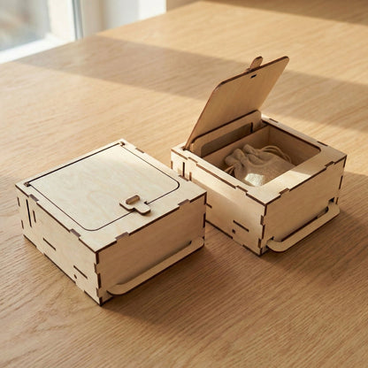 Two square laser cut plywood boxes on a wooden table. One is closed with a small latch; the other is open via a lever mechanism, revealing a small linen drawstring pouch inside. Features clean finger-joint edges and natural wood grain.