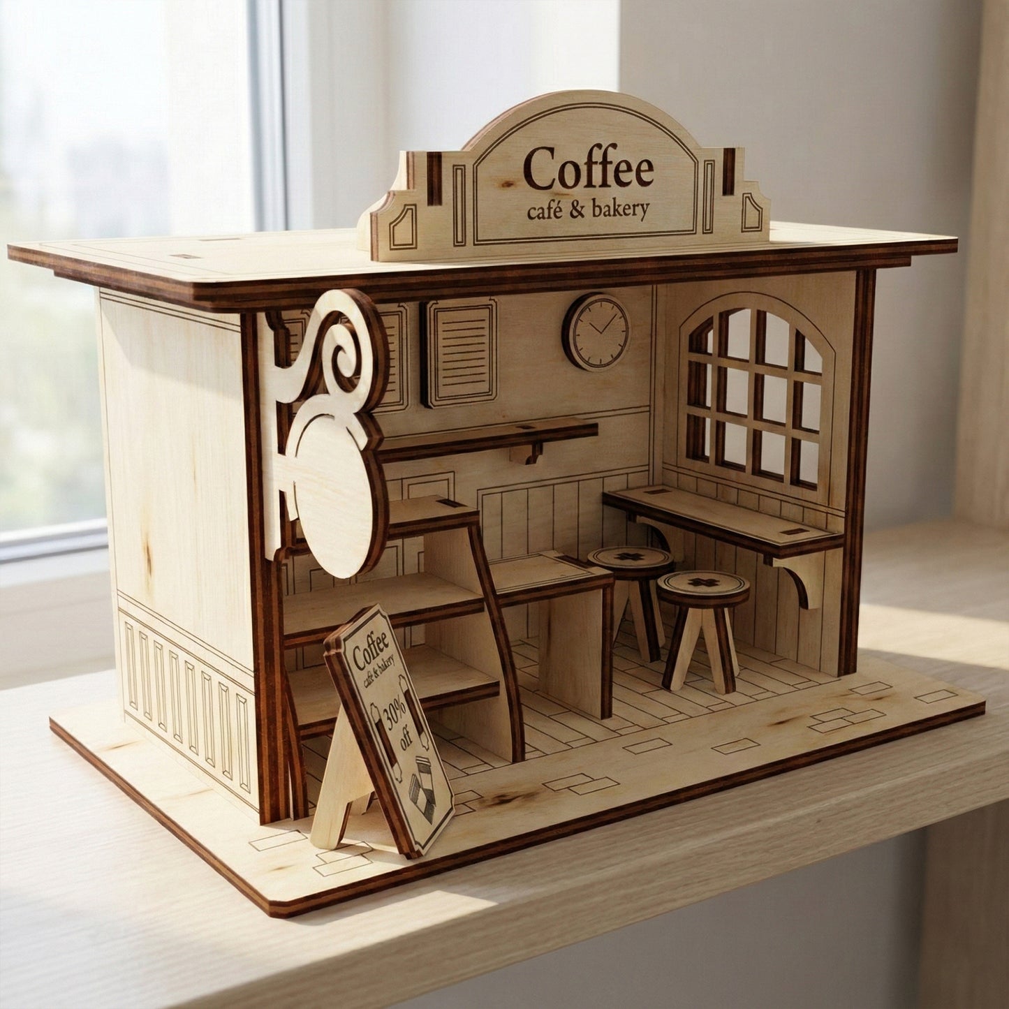 Side view of a miniature plywood cafe kit. The laser cut wooden structure sits on a light wood table, showing a window with a lattice frame, a wall clock detail, and tiny furniture. Natural sunlight highlights the smooth wood grain and dark edges.