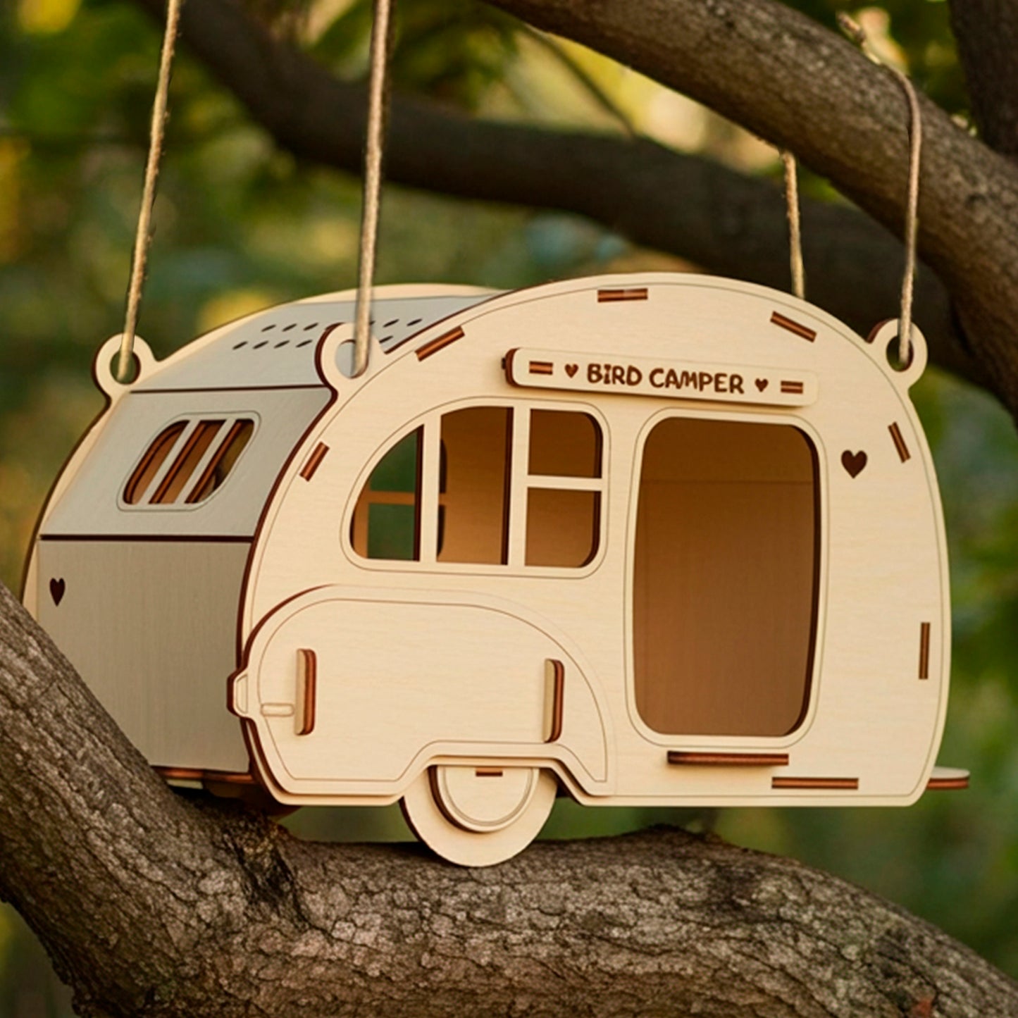 Laser cut plywood wooden birdhouse shaped like a retro camper trailer, hanging on a tree branch in a forest setting, surrounded by warm natural sunlight and soft green bokeh