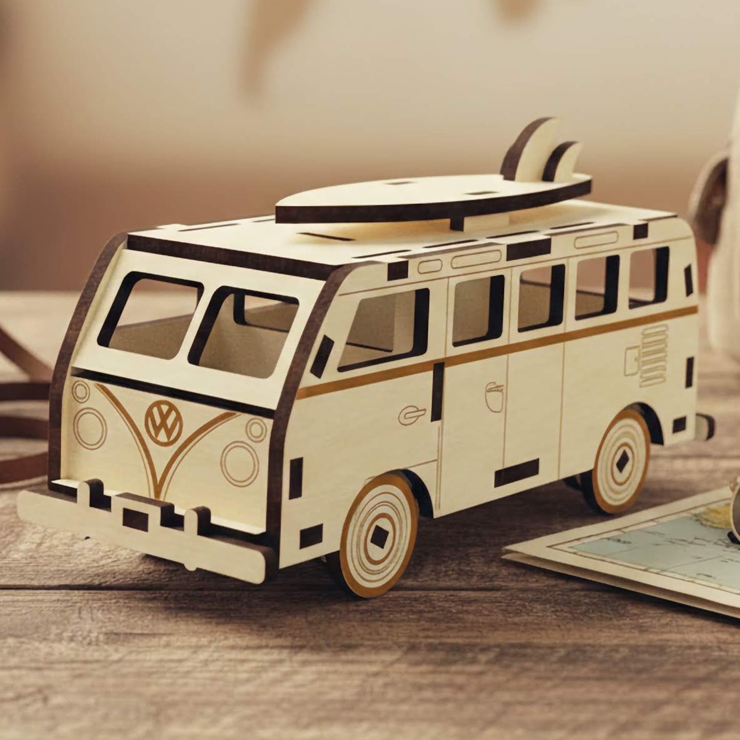Laser cut wooden plywood camper van miniature with a surfboard on top. Vintage style plywood model with engraved logo, resting on a wooden table next to a camera, map, and backpack. Detailed 3D wood puzzle for travel lovers.