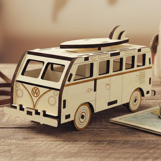 Laser cut wooden plywood camper van miniature with a surfboard on top. Vintage style plywood model with engraved logo, resting on a wooden table next to a camera, map, and backpack. Detailed 3D wood puzzle for travel lovers.