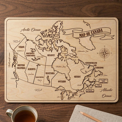 Laser cut plywood map of Canada with engraved province borders, city names, and oceans. Features a decorative banner and compass rose on natural light wood. Desktop display shown next to a cup of tea and pencil for size reference.