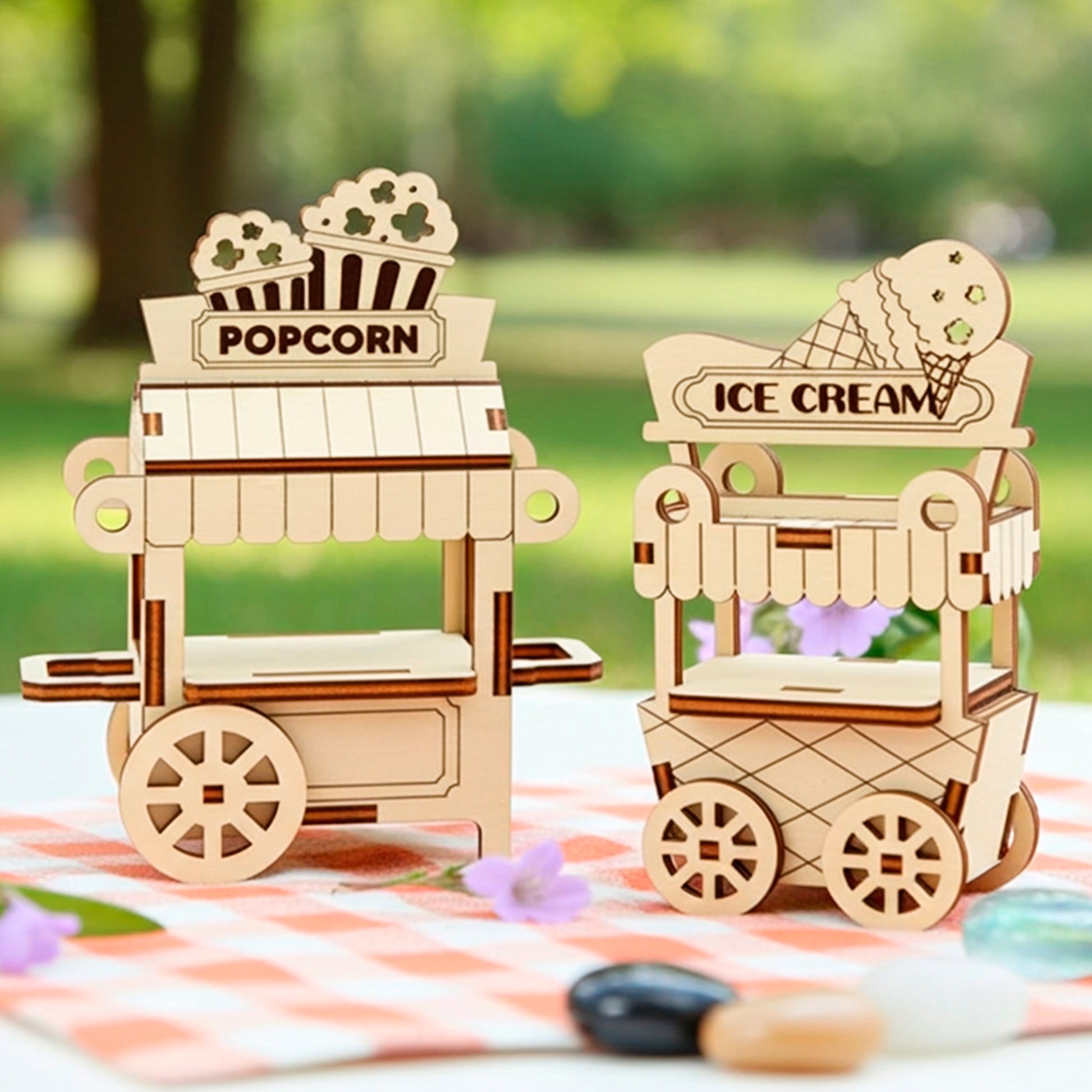 Two small, light-colored laser cut wooden plywood miniature food carts—one labeled "POPCORN" and the other "ICE CREAM"—displaying respective foods, set on a checkered tablecloth outdoors in a sunny park setting.